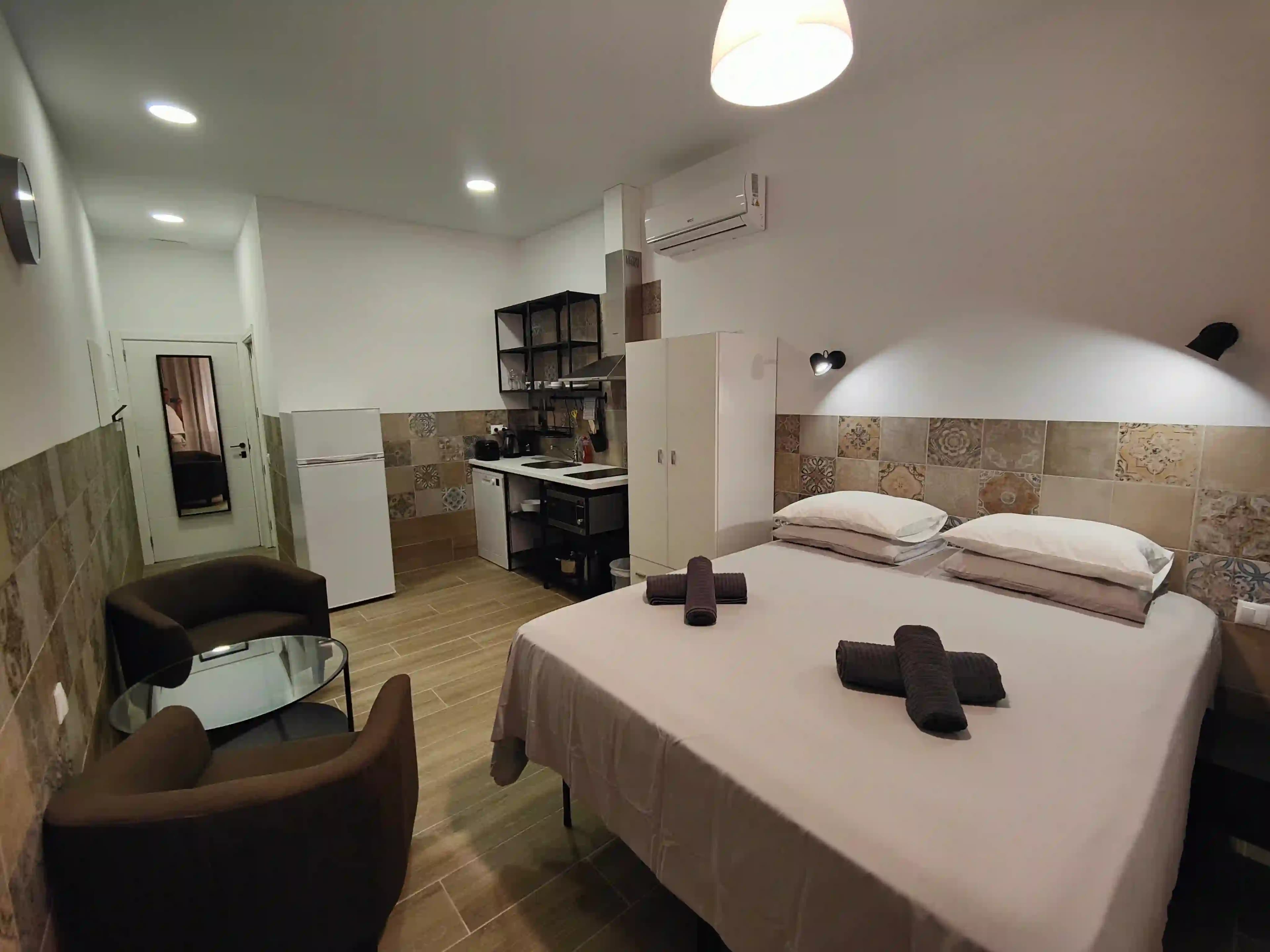 A double bed in a studio at Hotel Apartamentos Pueblo Mar, with two armchairs, a glass coffee table and the kitchenette behind
